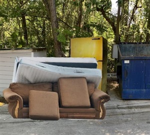 old sofa, mattress and fridge set next to blue trash dumpster