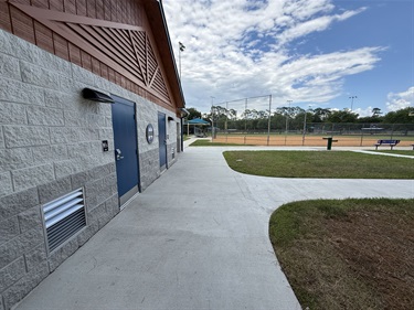 Exterior view of a park facility showing a restroom building, a curved concrete pathway, and a baseball field in the background.
